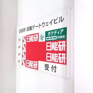 Building directory sign mounted on a wall, showing floor levels (2F–5F) with red panels and a green panel containing Japanese text.