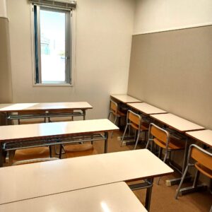 Empty classroom with rows of beige desks and wooden chairs, sunlight coming through a window on the left.