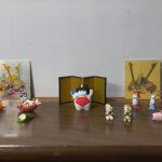 Row of colorful Japanese hina dolls and fish ornaments on a wooden shelf, with gold decorative boxes in the back.