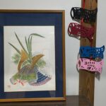 Framed botanical painting with green leaves and orange accents on a dark blue mat, mounted on a wall beside a vertical wooden post with colorful paper masks; the masks are black, red, pink, blue.