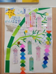 Colorful classroom poster with handwritten Japanese text, green paper strips, star shapes, and vertical wish strips featuring pink bear stickers for a summer goals display.