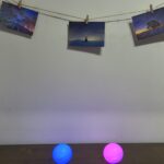 Three landscape photos hung on a string with clothespins against a plain wall, with blue and pink glowing balls on a dark table in the foreground.