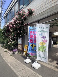 Storefront with colorful banners advertising classes, pink roses climbing the wall, and planters along the sidewalk.