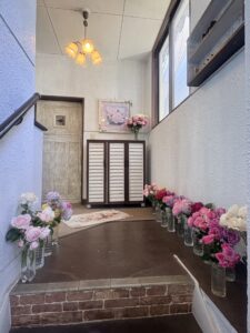 Narrow entry hallway lined with glass vases of pink and white roses on both sides, a wooden cabinet, and a floral painting above it.
