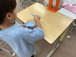 Child in a light blue shirt sits at a classroom desk, holding a blue pencil or tool over a blue plastic tray with a storage box and papers nearby.