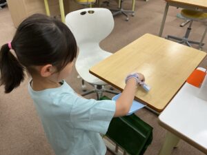 Young student in a light blue shirt wipes down a classroom desk with a cloth, at a study table in a clutter-free classroom.