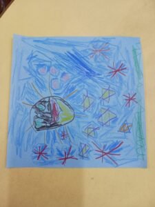 Child's crayon drawing on blue paper featuring a round globe with colored sections and orange rays, surrounded by red starbursts.