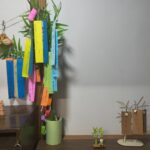 Bamboo branch with colorful paper Tanzaku strips hanging indoors, beside small clay figurines on a wooden table.