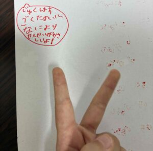 Hand making a peace sign in front of a white sheet with a red circular stamp containing handwritten red Japanese text inside the circle.