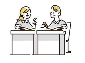 Two students sit at separate desks in a classroom, reading open books and talking to each other.