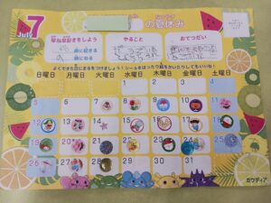 Bright yellow July calendar page with numbered days, sticker icons on several dates, and cartoon characters along the bottom edge, decorative fruits on the sides.