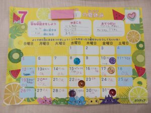 Bright yellow July calendar page with fruit decorations and cute characters along the bottom, featuring date boxes and handwritten notes in red and blue ink.